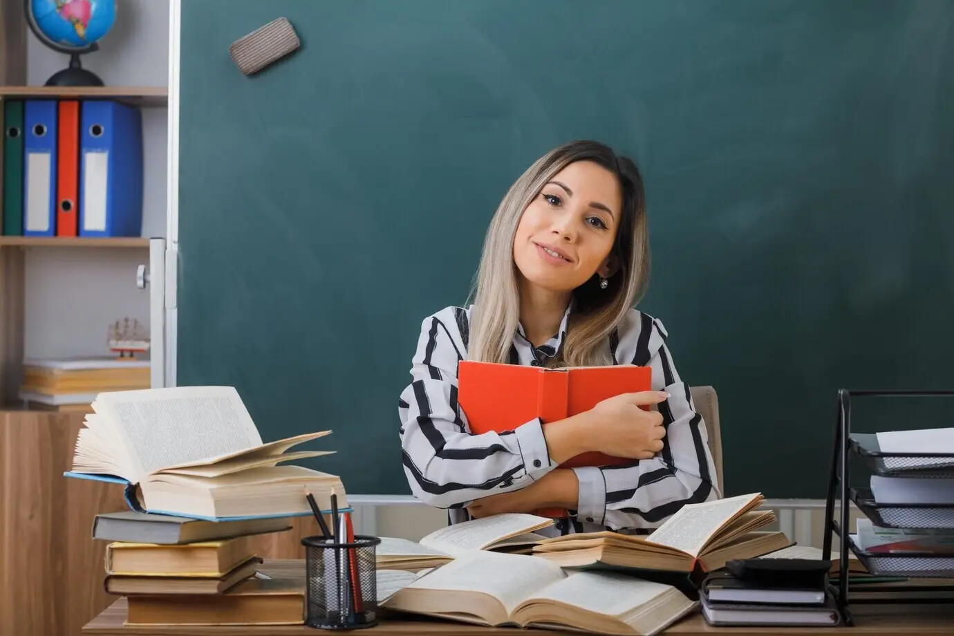 Eine junge Lehrerin sitzt im Klassenzimmer vor der Tafel an einem Schultisch, hält ein Buch, blickt in die Kamera und lächelt freundlich, glücklich und positiv.